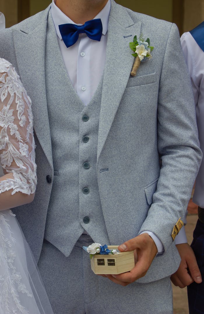 heros-img Close-up of an elegant groom wearing a gray suit and holding a wedding ring box.